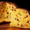 Panettone entero y una cuña cortada mostrando la miga amarilla con pasas y fruta confitada, sobre madera y fondo oscuro