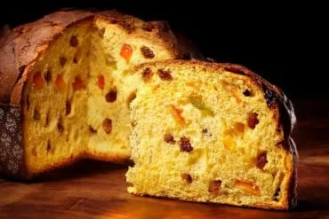 Panettone entero y una cuña cortada mostrando la miga amarilla con pasas y fruta confitada, sobre madera y fondo oscuro
