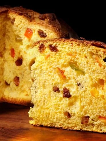 Panettone entero y una cuña cortada mostrando la miga amarilla con pasas y fruta confitada, sobre madera y fondo oscuro