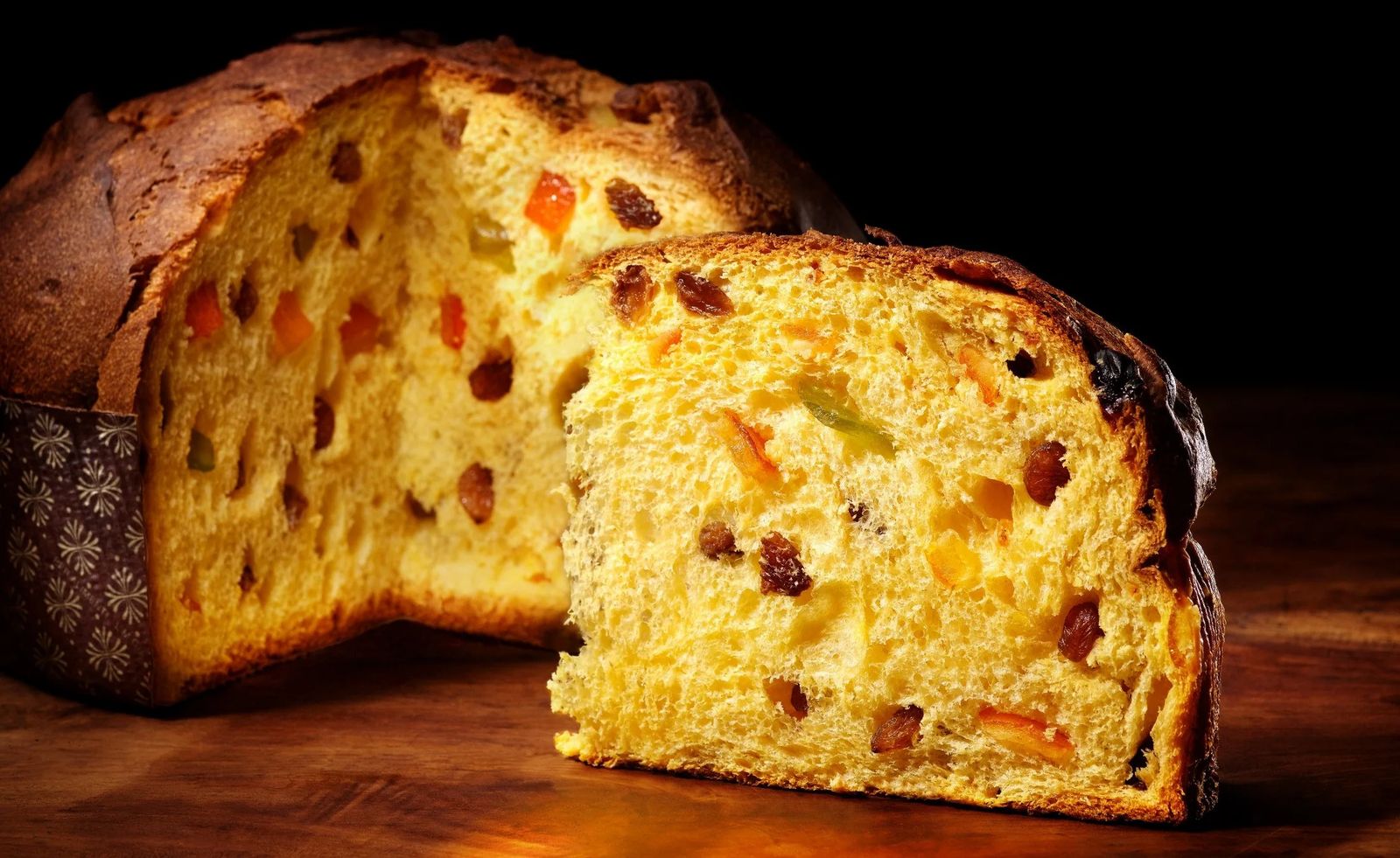 Panettone entero y una cuña cortada mostrando la miga amarilla con pasas y fruta confitada, sobre madera y fondo oscuro