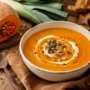 Crema de calabaza casera de color naranja intenso con una espiral de nata, pipas de calabaza tostadas y pimienta, servida en un cuenco rústico junto a una calabaza y un puerro