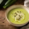 Crema de calabacín con queso de color verde suave, servida en un cuenco con aceite de oliva, virutas de queso, semillas de calabaza y pimienta, junto a calabacines frescos