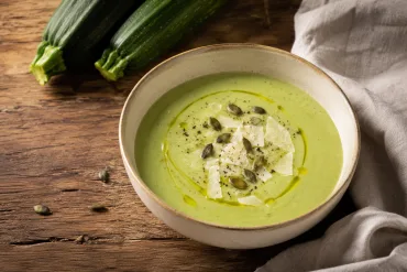 Crema de calabacín con queso de color verde suave, servida en un cuenco con aceite de oliva, virutas de queso, semillas de calabaza y pimienta, junto a calabacines frescos