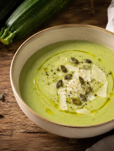Crema de calabacín con queso de color verde suave, servida en un cuenco con aceite de oliva, virutas de queso, semillas de calabaza y pimienta, junto a calabacines frescos