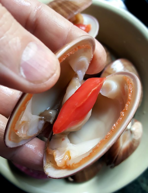 Una vez abierta, realizamos un corte en la parte más gruesa de la almeja para extraerla sus intestinos. Lavamos bajo un hilo de agua del grifo. Despegamos la carne de ambas conchas, quitamos una telilla de color marrón clarito y recolocamos la carne sobre una de las conchas y descartamos la otra. Colocamos la almeja limpia en el cuenco y limpiamos el resto de la misma manera.