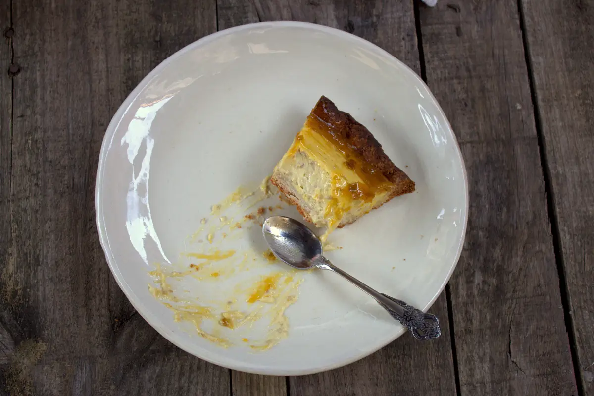 Media ración de tarta de queso y mango en un plato con restos y una cucharilla usada, con las capas de crema y mermelada a la vista