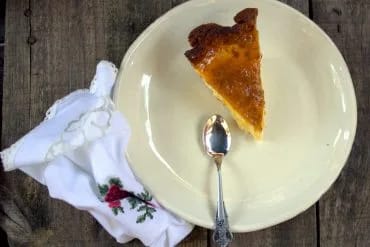 Ración entera de tarta de queso y mango con la mermelada brillante por encima, sobre un plato claro con una servilleta bordada