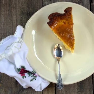 Ración entera de tarta de queso y mango con la mermelada brillante por encima, sobre un plato claro con una servilleta bordada