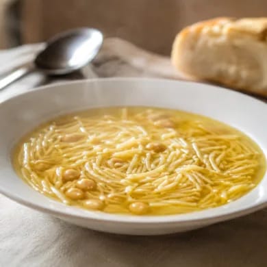 Caldo de cocido casero en plato hondo blanco, dorado y con fideos finos, con pan al fondo