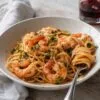 Plato hondo de espaguetis con langostinos en salsa de tomate y vermut, con perejil picado y una copa de vermut rojo al fondo